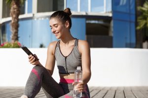 Outdoor shot of happy fitness woman looking at mobile phone app, sitting on wooden floor and drinking water